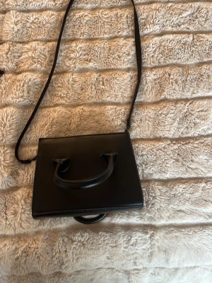 Svart axelväskaka i skinnimitation - Snygg svart axelv infolook med stilren, fyrkant det och både handtag och lång axelrem shaped. Väskan har ingen dragkedja upptill, det är en  minimalistisk design som passar till många outfits. Perfekt storlek för det viktigaste och enkel att bära crossbody.