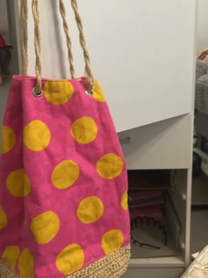 Rosa och gul prickig Strandväska - Färgglad axelväska i rosa tyg med stora gula prickar och grova rep som axelband. Väskan har en botten i flätad naturfiber som ger en somrig känsla. Perfekt för dig som vill sticka ut med en lekfull och unik accessoar.