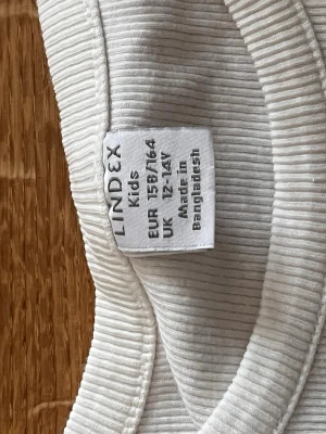 Vit ribbad linnetopp från Lindex - Säljer en enkel och stilren vit ribbad linnetopp från Lindex. Toppen är ärmlös och har en rund halsringning. Perfekt till sommaren och lätt att matcha med olika outfits.