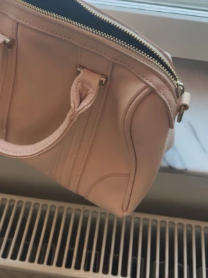 Ljusbeige handväska i skinnimitation - Snygg ljusbeige handväska i skinnimitation med guldfärgad dragkedja och dubbla handtag. Väskan har en stilren design med dekorativa sömmar och metallfästen. Perfekt storlek för det viktigaste och enkel att matcha till olika outfits.