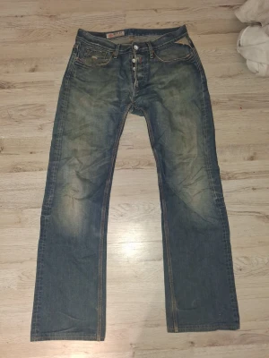 Replay blå jeans med raka ben - Säljer ett par klassiska blå jeans från Replay med raka ben och normal passform. Jeansen har slitna detaljer, knappar i gylfen och tydliga Replay-loggor på både fram- och baksida. Materialet är robust denim med snygg tvättad look och kontrastsömmar.  Finns ett litet hål i mellan bena men inga problem att sy igen. W32 L 36
