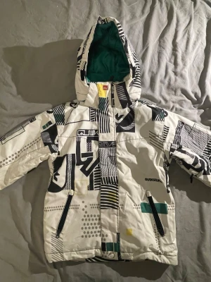 Skidjacka Quiksilver - Säljer en skidjacka från Quiksilver i storlek 10. Jackan är vit med svart, grått och grönt grafiskt mönster, har huva och grönt foder. Två dragkedjefickor framtill och vattenavvisande material. Perfekt för skidor och snowboard.