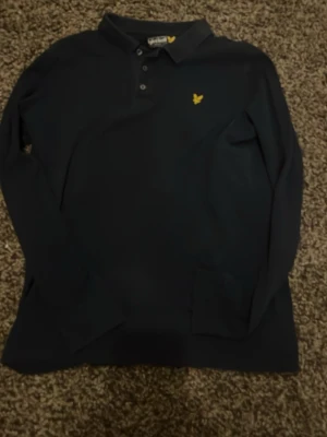 Mörkblå långärmad piké från Lyle & Scott - Mörkblå långärmad pikétröja från Lyle & Scott med klassisk krage och tre knappar framtill. Bröstet pryds av den ikoniska gula loggan. Storlek xs. 