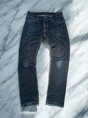 Mörkblå jeansbyxor från Nudie Jeans - Säljer ett par mörkblå jeansbyxor från Nudie Jeans med klassisk femficksdesign och kontrastsömmar. Jeansen har rak passform och är tillverkade i slitstarkt denimtyg av bomull. Snygga detaljer som nitar och Nudie-logga på bakfickan.