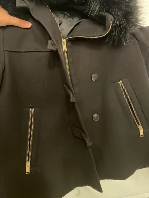 Svart kappa med fuskpäls och dragkedjor - Säljer en svart kappa med stor krage i fuskpäls och snygga guldfärgade dragkedjor på fickorna. Jackan har både knappar och dragkedja framtill för extra stil och värme. Perfekt för dig som vill ha en klassisk men ändå trendig look.