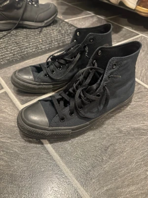 Converse - Säljer ett par helsvarta höga sneakers med klassisk rund tå och snörning. Skorna är i canvas med gummisula och har en stilren, enkel design som passar till det mesta. Perfekta för dig som gillar en clean och tidlös look.