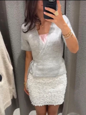 Ljusgrå omlottkofta med knyt - Superfin ljusgrå omlottkofta med kort ärm och knyt i sidan. Koftan har v-ringning och är stickad i ett mjukt material som känns skönt mot huden. Perfekt att styla med både kjol och jeans för en chill look.