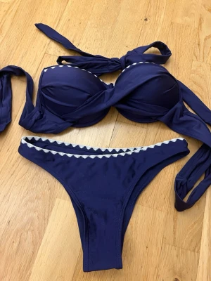 Mörkblå bikini med vit kant, står strl s i både topp och underdel - Snygg mörkblå bikini med vit sicksackkant längs både överdel och underdel. Överdelen har vadderade kupor och knytband för att justera passformen. 