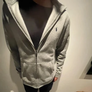 Grå hoodie från Polo Ralph Lauren - Grå hoodie från Polo Ralph Lauren med dragkedja och huva. Klassisk design med broderad logga på bröstet, två fickor framtill och mjukt material. Perfekt för chill dagar eller när du vill ha en clean och enkel look.