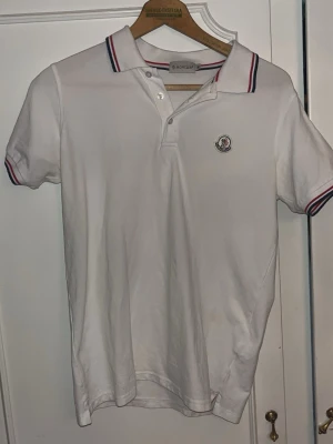 Vit Moncler piké med rödblå detaljer - Klassisk vit pikétröja från Moncler med korta ärmar och krage. Tröjan har rödblå ränder på krage och ärmslut samt Moncler-logga broderad på bröstet. Stängs med två vita knappar framtill. Perfekt till en clean och sportig look. Storlek M, passar S
