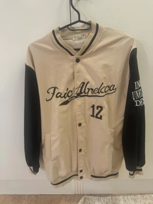 Beige och svart bomberjacka CH SE - Snygg bomberjacka i beige med svarta ärmar och broderade detaljer. Jackan har tryckknappar, ribbade muddar och coola broderier på bröstet och ena ärmen. Märkt med 'Faic Abnelca' och siffran 12 framtill. Perfekt för dig som gillar streetstyle. Köpt på beyond retro i befintligt skick (se bild 3 på lösa sömmar i brodyren), enbart använd fåtal gånger av mig! 