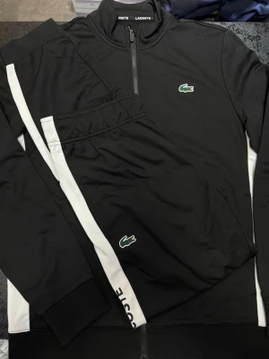 Svarta mjukisbyxor från Lacoste - Svarta mjukisbyxor från Lacoste med vit sidoremsa och klassisk grön krokodillogga. Byxorna har elastisk midja och är gjorda i mjukt syntetmaterial, perfekta för en sportig och avslappnad look.