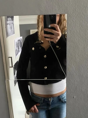 Svart kofta med guldknappar - Säljer en svart, croppad kofta med långa ärmar och snygga guldknappar framtill. Koftan har en klassisk krage och är i ett mjukt stickat material som känns både stilrent och trendigt. Perfekt att matcha med jeans eller kjol för en cool look.