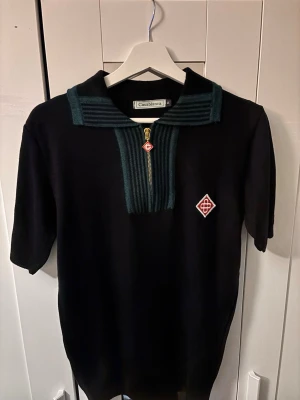 Svart topp Casablanca med dragkedja - Säljer en svart topp från Casablanca med gröna ribbade detaljer vid kragen och bröstet. Toppen har korta ärmar, guldfärgad dragkedja framtill och en broderad logga i rött och vitt på bröstet.