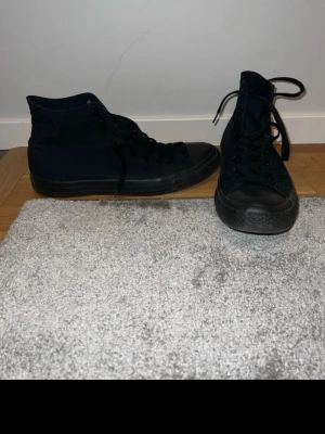 Svarta Converse Chuck Taylor All Star Unisex - Säljer ett par klassiska svarta Converse Chuck Taylor All Star high tops. Skorna har svart canvas, svart gummitå och svart sula. Ikonisk Converse-logga på sidan och snörning hela vägen upp. Perfekta till jeans eller shorts för en clean streetstil.