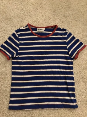 Randig t-shirt från Weekday, blå/vit - Säljer en randig t-shirt från Weekday i storlek S. T-shirten har blå och vita horisontella ränder med kontrasterande röd kant vid halsringning och ärmslut. Tillverkad i mjuk bomull och har en klassisk, avslappnad passform. Perfekt för dig som vill ha en chill vardag.