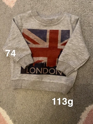 Grå sweatshirt med London-tryck - Grå sweatshirt med stort tryck av brittiska flaggan och texten 'London' framtill. Tröjan har ribbade muddar vid ärmslut och nederkant samt rund hals. Perfekt för dig som gillar coola prints och streetstyle.🌸 Fint använt skick. Storlek 74