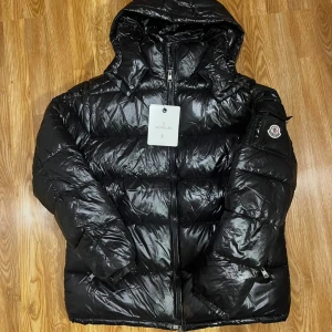 Svart Moncler pufferjacka med huva - Svart pufferjacka från Moncler i storlek L. Jackan har en glansig finish, huva, dragkedja framtill och Moncler-logga på ärmen. Fylld med dun och har praktiska fickor. Perfekt för kalla dagar och har en cool, modern look.