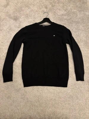 Svart stickad tröja från J.Lindeberg - Svart stickad tröja från J.Lindeberg med diskret logga broderad på bröstet. Storlek M, men sitter mer som S nästintill XS. Tröjan är i bra skick förutom det lilla hålet på sista bilden som man verkligen inte tänker på. Perfekt för dig som gillar stilrena och klassiska plagg med enkel design. Skriv vid minsta fråga eller fundering!