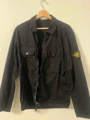 Svart overshirt från Stone Island - Säljer en svart overshirt från Stone Island med klassisk patch på ärmen. Jackan har två bröstfickor med knappar och dragkedja framtill. Perfekt för dig som gillar streetwear och vill ha en stilren look. Storlek S men passar M