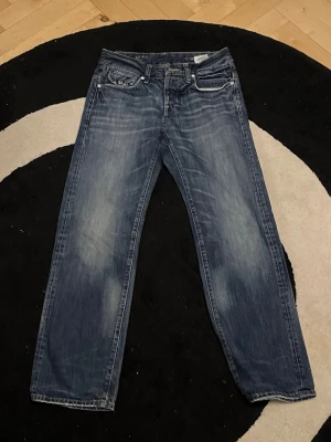 G-Star Morris Low Straight jeans - Säljer ett par blå G-Star Morris Low Straight jeans med klassisk femficksdesign och snygga slitningar. Jeansen har låg midja och raka ben, tillverkade i slitstark denim. Perfekta för dig som gillar en avslappnad och tidlös look.