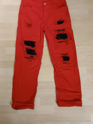 Röda ripped jeansbyxor  från Temu XL - Säljer ett par röda jeansbyxor i storlek XL med flera stora slitningar framtill och svarta detaljer under hålen. Byxorna har klassisk femficksdesign och är tillverkade i stretchig bomullsmix för skön passform. Från Temu!