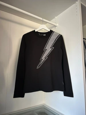 Svart tröja med blixt och nitar från Love Moschino - Cool svart långärmad tröja från Love Modchino, köpt på zalando, med en stor silverfärgad blixt på framsidan, dekorerad med nitar längs kanten. Tröjan har rund hals och är i mjukt material, perfekt för dig som vill sticka ut lite extra.