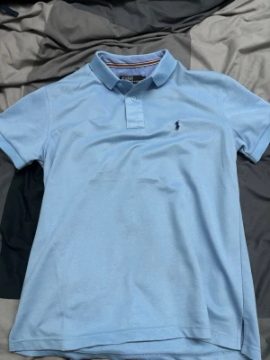 Ljusblå pikétröja Polo Ralph Lauren S - Klassisk ljusblå pikétröja från Polo Ralph Lauren i storlek S. Tröjan har korta ärmar, krage och knäppning framtill samt den ikoniska broderade loggan på bröstet. Tillverkad i bomull för en skön och luftig känsla.