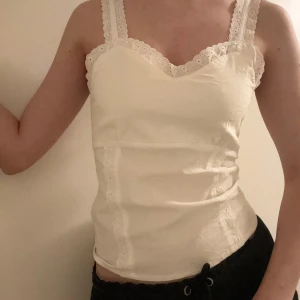 Vit spetskantad korsett från Wear It - Säljer en söt vit korsett från Wear It med breda axelband och spetsdetaljer längs kanterna. Toppen har v-ringad halsringning, hakknäppning bak och dekorativa sömmar framtill. Perfekt för dig som gillar romantisk och feminin stil.