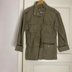 Olivgrön parkajacka från Lindex - Säljer en olivgrön parkajacka från Lindex med fyra stora fickor framtill, axelklaffar och hög krage med tryckknappar. Känns som en jeans jacka . Passar Xs och S