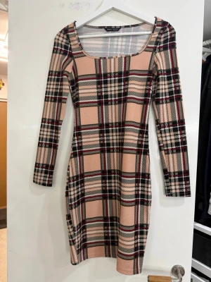 Rutig beige och svart kortklänning - Säljer en snygg kortklänning från Shein med rutigt mönster i beige, svart, vitt och röda detaljer. Klänningen har lång ärm och fyrkantig ringning. Materialet är stretchigt och känns mjukt mot huden. Perfekt för dig som gillar trendiga och bekväma plagg.