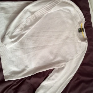 Vit stickad tröja från BOSS, M - Vit stickad tröja från BOSS i storlek M. Tröjan har rund halsringning med en gul rand på insidan av kragen och diskreta ränder på ärmarna. Perfekt för dig som gillar stilren och klassisk design.