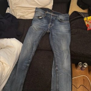 Blå jeans från Nudie Jeans Co, W32 L32 - Säljer ett par klassiska blå jeans från Nudie Jeans Co i storlek W32 L32. Jeansen har snygga slitningar, raka ben och femficksmodell. De är tillverkade i mjukt denimtyg med orangea sömmar och har Nudie-logga på bakfickan.