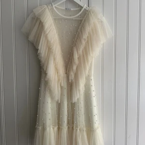 Vit Ida Sjöstedt klänning - Vit/cream klänning från Ida Sjöstedt, modell Nathalie med volangdetaljer, pärlor och djup v-ringning. Klänningen är i tunn tyll med knytband i midjan. Perfekt för studenten! Endast använd en gång. Köpt för 4999 kr, pris kan diskuteras!