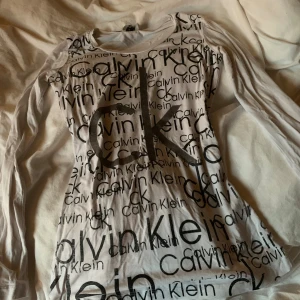 Vit långärmad Calvin Klein topp - Vit långärmad topp från Calvin Klein Jeans med svart logotyp- och textprint över hela framsidan. Slimmad passform och rund halsringning. Tillverkad i mjuk bomull, perfekt för en clean och trendig look.