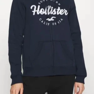Mörkblå Hollister hoodie med dragkedja - Säljer en mörkblå hoodie från Hollister med vit broderad logga på bröstet. Tröjan har huva med snörning, dragkedja framtill och en stor magficka. Perfekt för chill dagar och enkel att matcha med jeans eller mjukis. Är i storlek M men passar även mig i S lite oversize. Skriv om du har frågor eller vill ha fler bilder!🥰