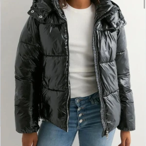 Svart glansig pufferjacka med huva - Säljer en svart glansig pufferjacka med huva och dragkedja framtill. Jackan har quiltad design, långa ärmar och praktiska fickor med knappstängning. I st S säljer då den inte passar! 
