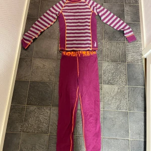 Randig merinoullströja lila/orange 160 - Långärmad underställströja i lila och grått med ränder och orange sömmar. Tillverkad i 70% merinoull och 30% syntet, perfekt för kalla dagar. Snygg sportig look med kontrastfärgade detaljer och normal passform.