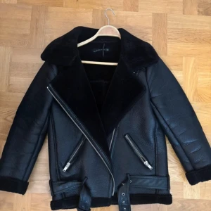 Svart pälsjacka med bälte och dragkedjor - Säljer en svart pälsjacka från Zara med bred krage, två sneda dragkedjefickor och bälte i midjan. Jackan har en oversized passform och är fodrad med mjuk pälsimitation. Perfekt för kalla dagar och har snygga detaljer som synliga sömmar och grova dragkedjor.