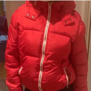 Röd pufferjacka med huva - Säljer en röd pufferjacka med huva och vita dragkedjor. Jackan har en kortare, lite boxig passform och två fickor framtill med dragkedja. Perfekt för kalla dagar när du vill hålla stilen och värmen.
