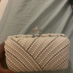 Pärlbeströdd vit kuvertväska med stras - Superfin kuvertväska täckt av vita pärlor och glittrande strasstenar. Väskan har ett elegant ringhandtag med stenar och silvriga detaljer. Perfekt accessoar för att lyfta din look med extra glamour. Insidan är fodrad i vitt tyg och väskan stängs med ett smidigt klicklås.