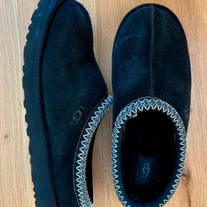 Svarta UGG Tasman mockatofflor strlk 44 - Svarta UGG Tasman tofflor i mocka med broderad kant i vitt och svart. Använda förra vintern, nu urvuxna. Inga defekter, gott använt skick. Kommer i originalboxen. 