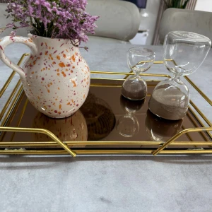 Dekorativ bricka - Bilderna visar en dekorativ bricka med guldfärgad metallram och spegelglasbotten, samt en keramikkanna med blommor och två timglas. Brickan har modern design och ser ut att vara i mycket gott skick utan synligt slitage. Perfekt för servering eller som elegant inredningsdetalj.