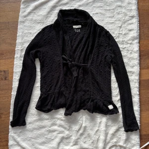 Svart kofta med knyt från Odd Molly storlek 4 - Supersöt svart kofta från Odd Molly i 100% bomull. Koftan har ett fint hålmönster, volangkant nertill och knytband framtill. Långärmad och perfekt för lager-på-lager-stil. Snygg och bekväm att slänga på sig när det blir lite kyligt. Perfekt skick