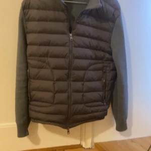 Moncler cardigan  - Snygg pufferjacka i mörkgrått och svart med quiltad framsida och mjuka grå ärmar och rygg. Jackan har hel dragkedja framtill och två fickor med dragkedja. Perfekt för kyliga dagar när du vill ha både stil och komfort.