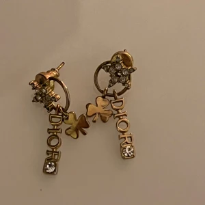 Guldiga örhängen  - Unika guldiga örhängen hängande fyrklöver, gnistrande kristaller och -logga. Örhängena har små stjärnor dekorerade med stenar och en exklusiv känsla som verkligen sticker ut.