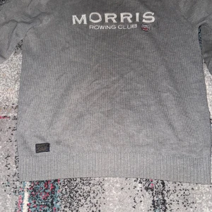 Grå stickad tröja från Morris - Snygg grå stickad tröja från Morris med broderad text 'Morris Rowing Club' och liten sköld på bröstet. Tröjan har rund hals, långa ärmar och ribbade muddar. Perfekt för dig som gillar klassisk och stilren look.