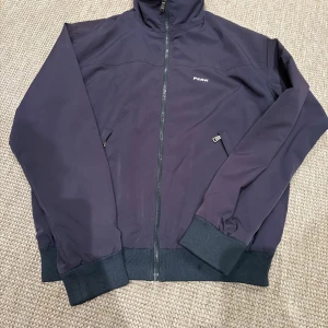 Mörkblå vindjacka Peak Performance L - Snygg mörkblå vindjacka från Peak Performance med dragkedja framtill och två fickor med dragkedja. Jackan har ribbade muddar vid ärmslut och midja, samt hög krage. Perfekt för dig som gillar stilren och sportig look.