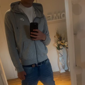Grå Nike hoodie med dragkedja - Säljer en stilren grå hoodie från Nike med hel dragkedja och huva. Tröjan har långa ärmar, två fickor framtill och klassisk Nike-logga på bröstet. Perfekt för chill dagar eller när du vill ha en sportig look.