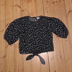 Blommig svart blus från Lindex - Supersöt svart blus från Lindex med små blå och vita blommor. Modellen har puffiga trekvartsärmar och en knytning framtill som ger en trendig look. Perfekt för dig som gillar romantiska detaljer och blommigt mönster.